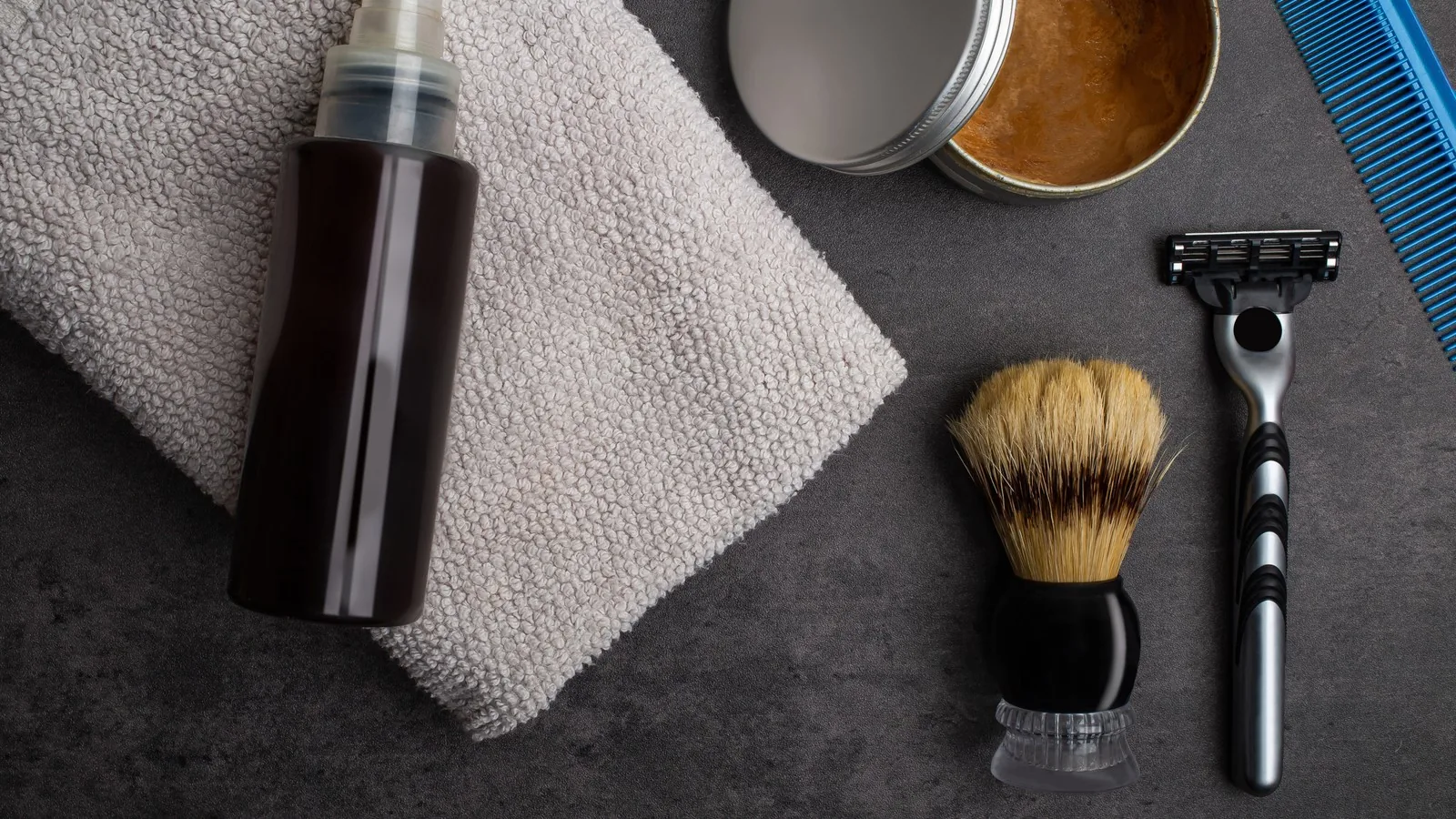An overhead flat lay of unbranded men's grooming items on dark slate: an amber spray bottle, a folded grey towel, an open tin of balm, a shaving brush, a razor and a comb.