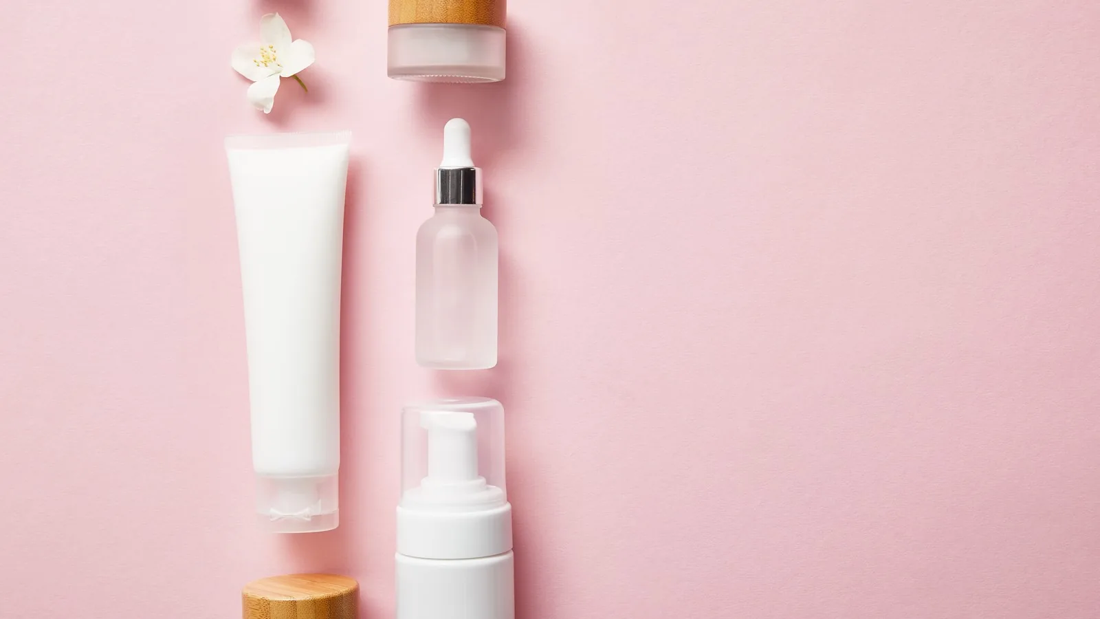 An overhead flat lay of unbranded white cosmetic containers on a soft pink background: a cream tube, a dropper bottle, a pump bottle and two lidded jars beside a jasmine flower.